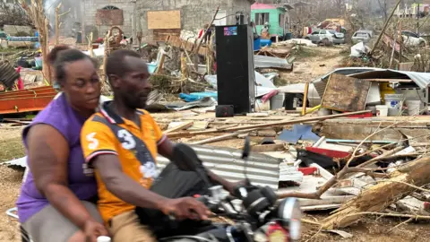 Brandon Drenon / BBC News Two people on a motorcyle ride through a town covered in storm debris including broken tree branches, parts of buildings and cars 