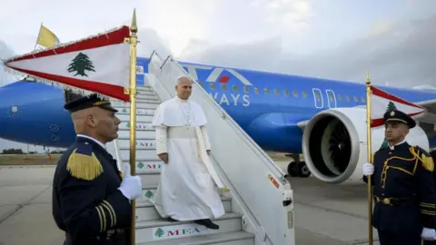 Getty Images A man in white religious robes and skullcap descends the steps of an airliner as soldiers in dress uniform stand guard with flags. 