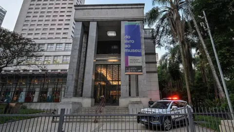 AFP via Getty Images A police car with flashing lights stands in  front of the main entrance to the Biblioteca Mario de Andrade in São Paulo. A uniformed police officer is standing guard next to the entrance. A banner hanging from the column at the left-hand side of the entrance advertises the exhibition "From book to museum".