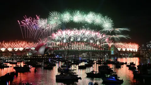 AFP via Getty Images Fireworks light up the midnight sky over Sydney Harbour Bridge and Sydney Opera House during New Year's Day celebrations in Australia. Boats are parked in the harbour facing the fireworks display.