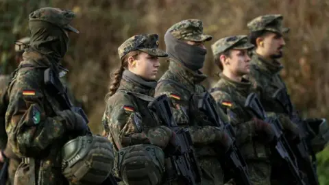 Reuters Male and female soldiers stand with guns in a field, wearing camouflage with the German flag stitched on their upper arms.