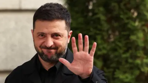 Reuters Ukrainian president Volodymyr Zelensky, a man with dark hair and beard, wearing a dark shirt, raises his left hand to the camera.