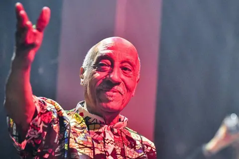 Mulatu Astatke Mulatu Asatke wearing a patterned shirt smiles and waves to the audience.