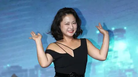 Getty Images South Korean comedian Park Na-Rae with a short black bob and a black dress waving against a blue background at a press event
