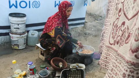 Reuters Displaced Palestinian woman Hanan Abu Taibah cooks food on a fire outside her  tent in Khan Younis, southern Gaza (18 December 2025)