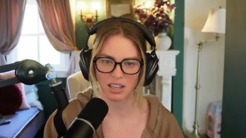 BBC A woman, QTCinderella, is sitting wearing black over-ear headphones and black glasses, with a brown, open hoodie. She is speaking into a black microphone. In the background is a lamp and a vase of flowers and a window with pink curtains. 