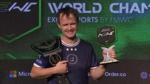 MEWC A man with short brown hair smiles, holding a wrestling style belt, and a trophy in hand. He is a navy-blue E-sports jersey with sponsors on the arms. Behind him is a display board, featuring Microsoft Excel World Championship, and the event sponsors listed at the bottom.