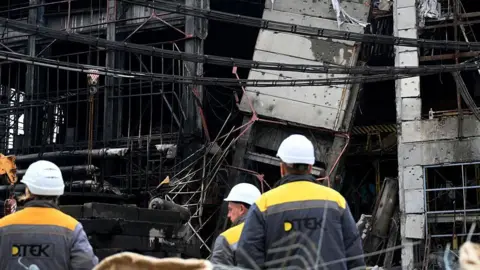 YURIY DYACHYSHYN/AFP Employees stand next to the building of a power plant of Ukrainian energy provider DTEK, which was heavily damaged during air attacks, at an undisclosed location on December 10, 2025, amid the Russian invasion of Ukraine