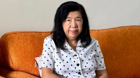 BBC Susanna Liew, wearing a white top with black polka dots, is seated on a brown sofa and looking straight at the camera
