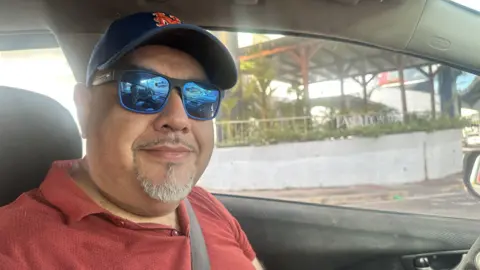 BBC A man in the drivers seat of a car, wearing a blue cap and blue sun glasses looking into the camera.