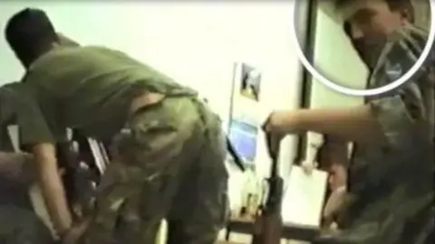 US Immigration and Customs Enforcement A photo released by US Immigration and Customs Enforcement, showing a man circled on the right-hand side holding what appears to be a gun. Another man on the left is bending over. Both are wearing what appears to be military clothing.