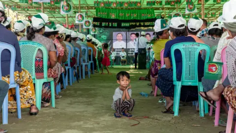 Jonathan Head/ BBC A child sitting on the ground in the aisle between two rows of chairs at a campaign rally in Mandalay. He is looking away from the stage, towards the camera.    
