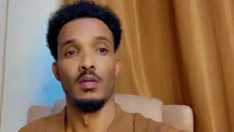 Mahad Mohamud A head and shoulders image of Mahad Mohamud, He is wearing a sandy brown shirt and a similar coloured curtain can be seen behind him.