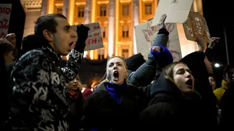AFP via Getty Images A crowd of protesters holding cardboard signs shouts outside a grand, lit-up building in Sofia at night.