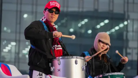 BBC/Hosu Lee A person in sunglasses, a red scarf and a red cap that says 'MAKE KOREA GREAT AGAIN' bangs a drum hanging from his front, while a woman beside him does the same