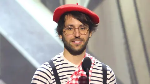 Picture Group A man in a red beret and striped t-shirt stands on stage behind two microphones as he delivers a speech.