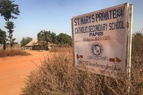 AFP via Getty Images A sign that reads St. Mary's Private Catholic Secondary School Papiri, Niger State, is positioned on the right with red dirt road and a building in the background surrounded by trees.