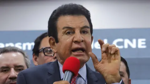 Reuters Presidential candidate Salvador Nasralla of Honduras' Liberal Party (PLH) gestures during a press conference. He wears a navy suit, blue short and red patterned tie. He is speaking into a bright red microphone