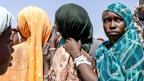 AFP via Getty Images The heads and shoulders of three women standing close together in a queue. They are wearing headscarves - one orange, one black and one turquoise and two are facing away from the camera and one is looking towards it. 