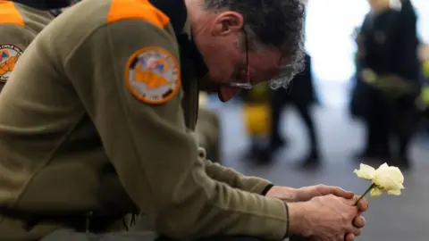 EPA A bespectacled member of the civil protection unit wearing a khaki uniform, squats as he holds a white rose with both hands, with his head lowered 