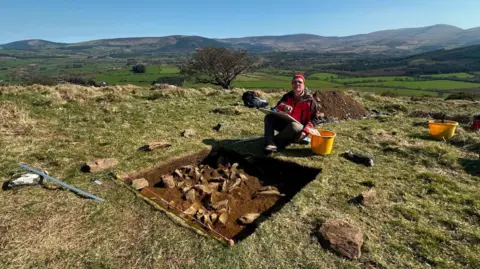Antiquity A man wearing a red soft shell jacket and red hat kneeling down in front of a hole during an excavation.