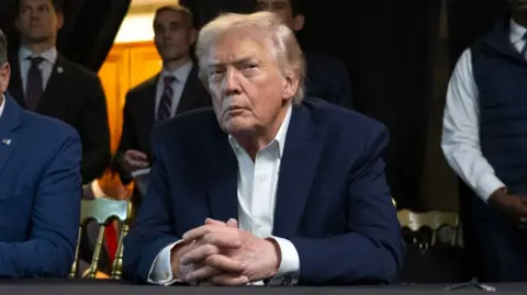 Getty Images Donald Trump sits with his hands interlocked on a desk during the raid of Caracas. He wears an open-neck white shirt and blue blazer.