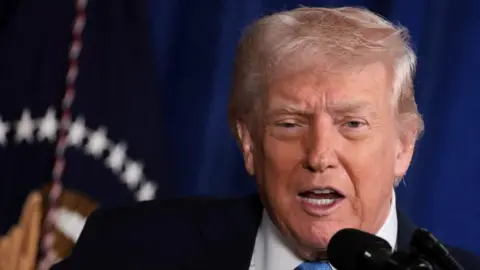 Reuters Donald Trump stands in front of a blue background and speaks at a microphone. He is wearing a black suit, white shirt and blue tie. 