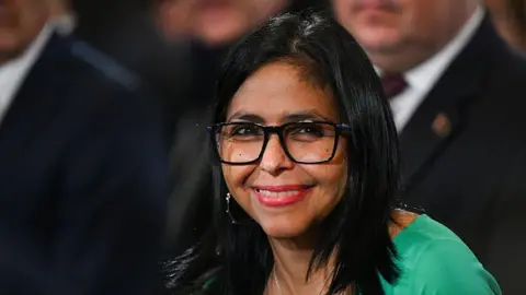Federico PARRA / AFP via Getty Images Venezuela's interim President Delcy Rodriguez smiles after a session of the National Assembly in Caracas on January 5, 2026. She is wearing black-rimmed glasses and silver earrings. Her dress is a lime green colour. 