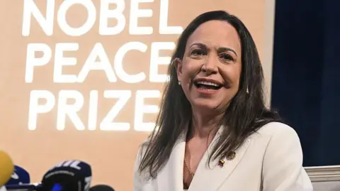Getty Images María Corina Machado wears a white suit and smiles in front of a sign that says "The Nobel Peace Prize".