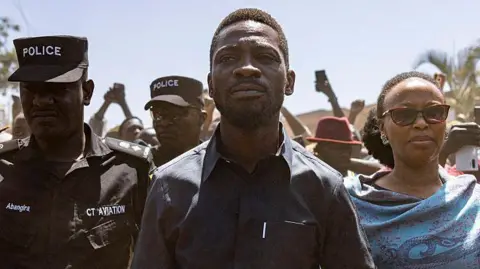 Getty Images Bobi Wine in a black shirt flanked by a police officer and his wife in a blue shawl on voting day.