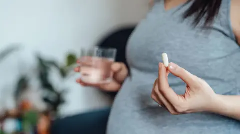 Getty Images A pregnant women wearing a grey top is visible from the chest down, and holds a white pill in her left hand and a glass of water in her right (slightly blurred)