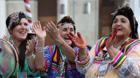 APP/NurPhoto via Getty Images Three women clap, wearing colourful clothing and jewelry