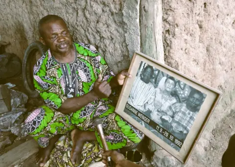Molara Wood Kasali Akangbe Ogun, dressed in a colourful green outfit, is interviewed while holding a frame with other members of the New Sacred Movement