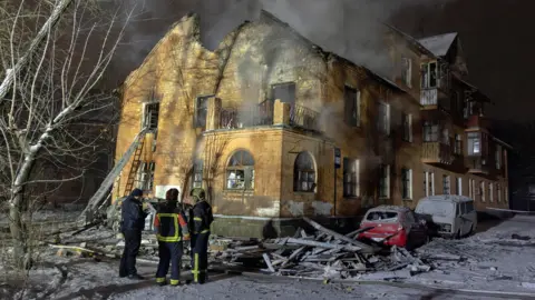 Reuters A smouldering residential building with a few firefighters standing out the front. There is lots of rubble around the building and covering a few cars that are parked out the front, and a bit of smoke coming from inside the building.
