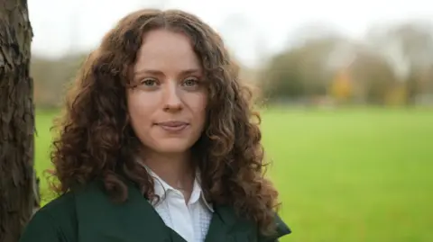 BBC Holly stares to camera. She is in focus with dark, long curly hair and is leaning against a tree-trunk while the background is out of focus but looks like a green field.