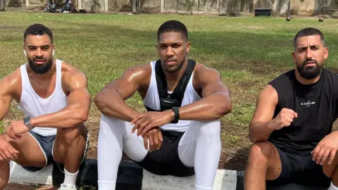Social media Anthony Joshua sits between Sina Ghami and Latif Ayodele as all wear training kit, with a grassy area behind the three men.
