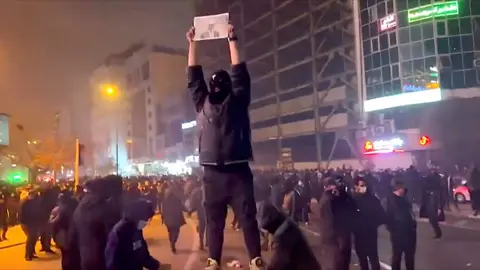 Public domain A masked protester stands on a platform above crowds at night in Kaj Square, in north-west Tehran. on 9 January 2026. They are holding a black and white photo above his head of the last shah's son, Reza Pahlavi. Light from buildings, streetlamps and shop fronts are illuminating the area. The protester's mask is black with a white mark on the front, with three holes cut out for their eyes, mouse and nose.