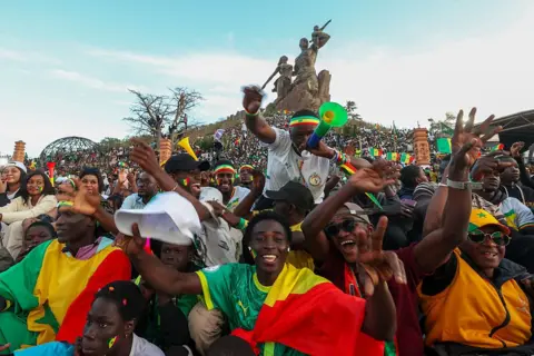 Andalou via Getty Images Senegalese fans wearing clothing and accessories in the colors of the national flag gather around African Renaissance Monument, chanting and singing to support their team.