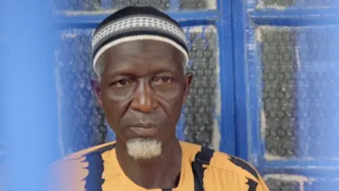 BBC Foday Musa, who has a white goatee, looks solemn. He wears a yellow and black top and white and black knitted skull cap and behind him is a window with blue painted frames.
