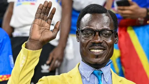 AFP/Getty Images Michel Nkuka Mboladinga dressed as Patrice Lumumba in a yellow suit jacket and blue cheque shirt and tie lifts his hand as if waving in the stands at Afcon.
