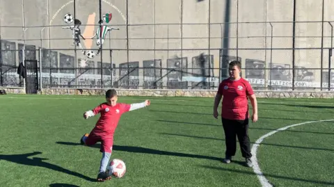 BBC Two 10-year-old boys play football together. Both are wearing red football shirts - one is about to kick the ball while the other watches. They are playing on a green pitch which is directly in front of a large concrete wall that towers over them. 