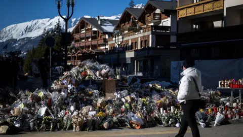 AFP via Getty Images A woman stands looking at piles of flowers, candles and messages at a makeshift memorial near Le Constellation bar in the luxury Alpine ski resort of Crans-Montana on 6 January. Behind the memorial are alpine-style wooden buildings with the mountains situated in the background.