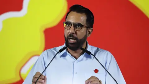 Getty Images Pritam Singh, wearing a light blue shirt and a red WP pin, gestures with his hands as he speaks at a lectern with microphones, against a red and yellow backdrop